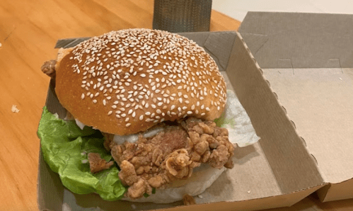 A sesame seed bun sandwich with crispy fried chicken, lettuce, and mayonnaise in a cardboard box on a wooden table.