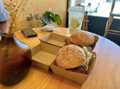 Two sesame seed burgers in takeout boxes on a wooden table, with a drink and a vase of dried flowers in the background.