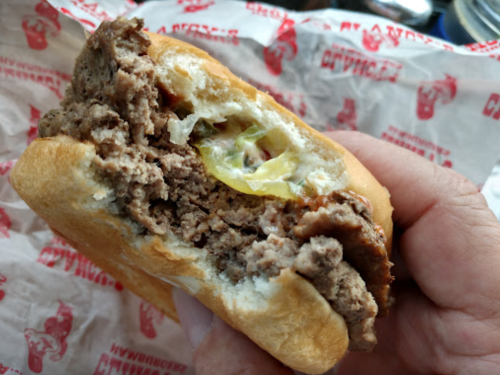 A hand holding a half-eaten burger with ground beef, pickles, and a soft bun, resting on red patterned paper.