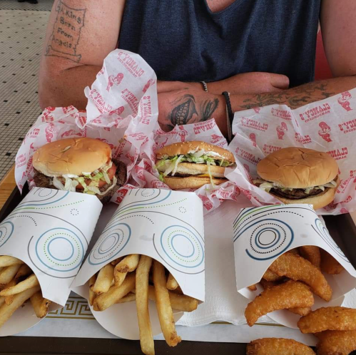 A person with tattoos sits at a table with three burgers and two servings of fries on a tray.