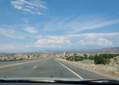 A wide, empty highway stretches into the distance under a blue sky with scattered clouds and distant mountains.