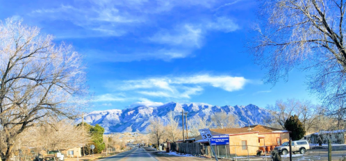 A scenic view of a snowy mountain range under a blue sky, with a road and buildings in the foreground.