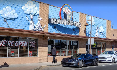 A storefront with a colorful mural, featuring a cow theme, and two parked cars in front. Sign reads "We're Open 7 AM to 9 PM."