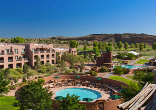 A scenic view of a resort with a circular pool, surrounded by greenery and mountains under a clear blue sky.