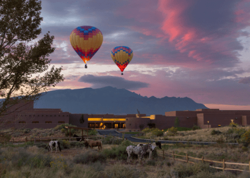 Two hot air balloons float above a landscape with cows, mountains, and a colorful sunset sky.