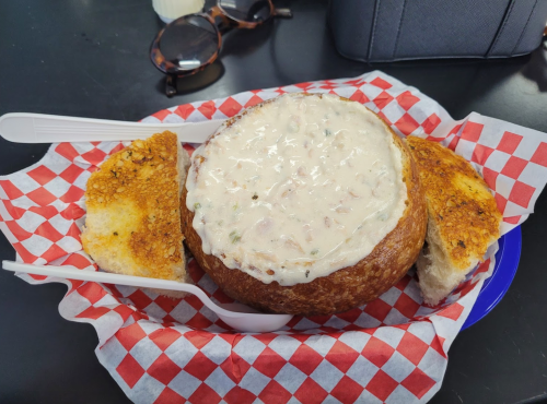 A bowl of clam chowder served in a bread bowl, accompanied by two pieces of toasted bread, on a checkered paper.