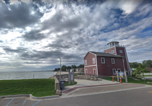 A red building by the water under a cloudy sky, with a grassy area and a paved path in the foreground.