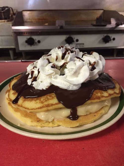 A stack of pancakes topped with whipped cream and chocolate sauce, with a creamy filling between the layers.