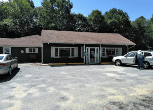 A single-story building with a brown roof, surrounded by trees, and two parked cars in a gravel lot.