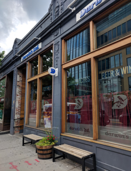Exterior of Darwin's Ltd. featuring large windows, a sign, and a bench with flower planters outside.