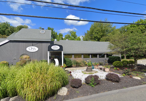 A gray building with a sign reading "CIMA," surrounded by greenery and a landscaped garden under a blue sky.