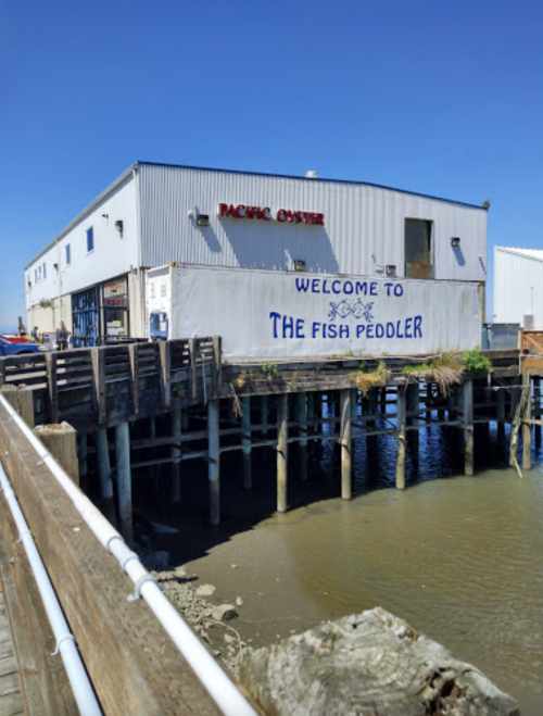 A waterfront building with a sign reading "Welcome to The Fish Peddler" and "Pacific Oyster" under a clear blue sky.