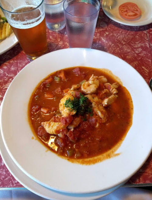 A bowl of seafood stew with shrimp and vegetables, garnished with parsley, next to a glass of beer.