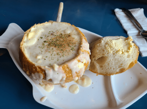 A bowl of creamy soup served in a bread bowl, garnished with herbs, alongside a piece of bread.