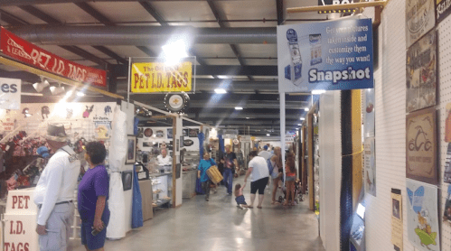 A bustling indoor market with various stalls, shoppers browsing, and signs for pet tags and photo services.