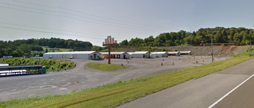 A wide view of a storage facility with several buildings and a large sign, surrounded by green hills and a clear sky.