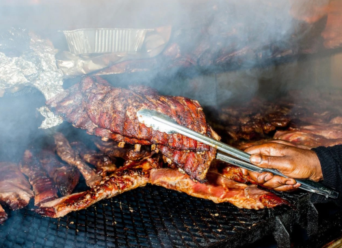 A hand using tongs to lift a rack of smoky, grilled ribs from a barbecue grill, surrounded by more meat.