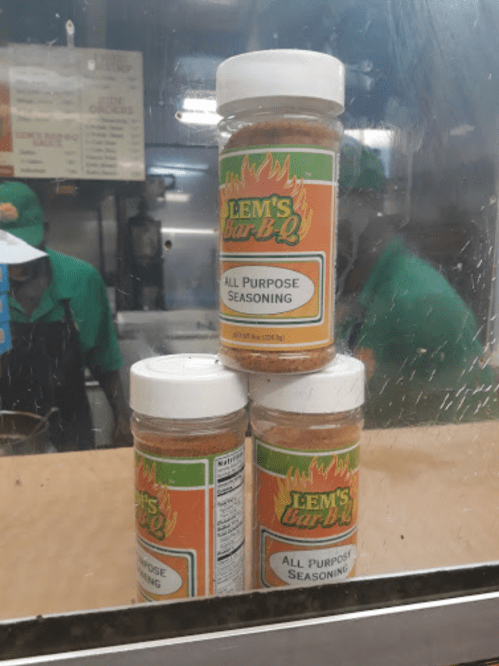 Three jars of Lem's Bar-B-Q All Purpose Seasoning stacked on a counter, with a blurred background of a kitchen.