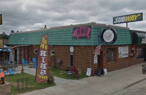 A rustic BBQ restaurant with a green roof, outdoor seating, and a Subway next door, featuring colorful signage.