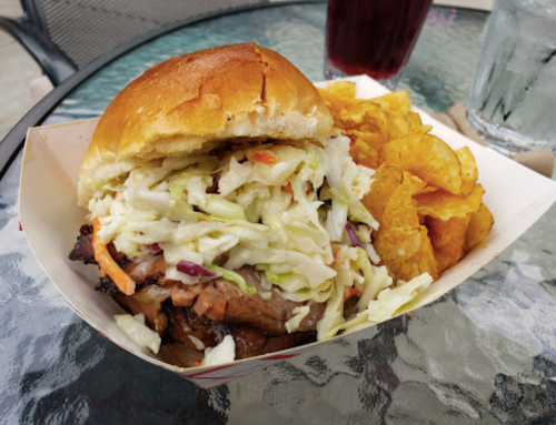 A close-up of a pulled pork sandwich topped with coleslaw, served with a side of potato chips and drinks in the background.
