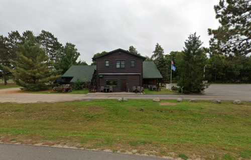 A two-story log cabin-style building with a green roof, surrounded by trees and a gravel parking area.