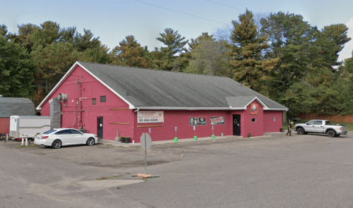 A red building with a gray roof, surrounded by trees, featuring a parking lot and signage for a business.