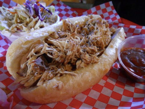 A pulled pork sandwich in a bun, served with coleslaw and a side of barbecue sauce in a red-checkered basket.