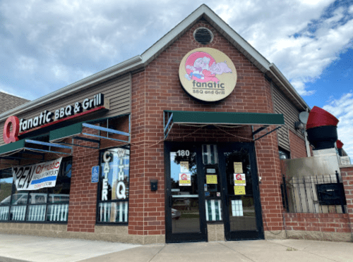 Exterior of Fanatic BBQ and Grill, featuring a brick facade, green awnings, and a large sign with a pig logo.