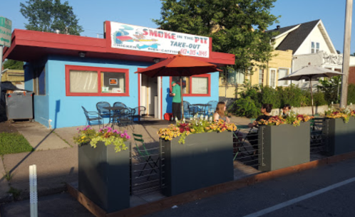 A colorful take-out restaurant with outdoor seating and flower planters, set in a sunny neighborhood.