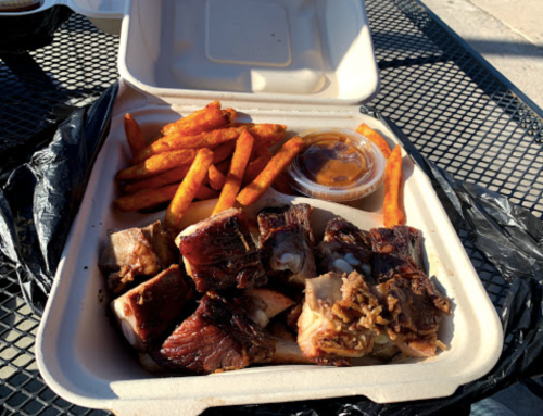 A takeout container with BBQ ribs, sweet potato fries, and a small cup of sauce on a black table.