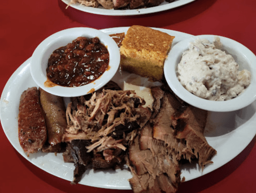 A plate of barbecue featuring pulled pork, brisket, sausage, baked beans, cornbread, and coleslaw on a red table.