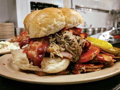 A hearty sandwich stacked with pulled pork, bacon, and pickles, served with a side of crispy potato wedges.