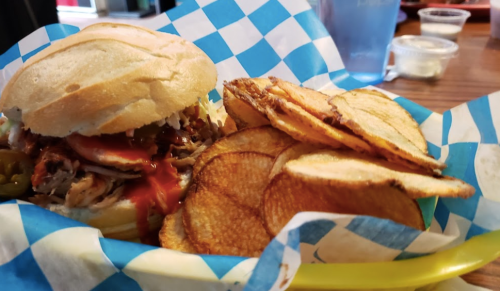 A pulled pork sandwich with jalapeños and barbecue sauce, served with crispy potato chips in a blue checkered basket.