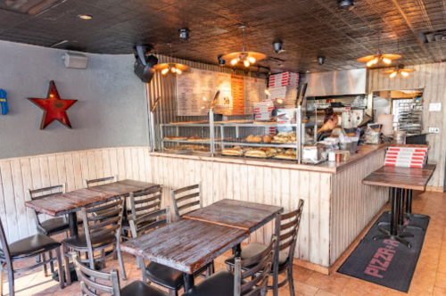 A cozy pizza shop interior with wooden tables, a counter displaying pizzas, and a staff member preparing food.