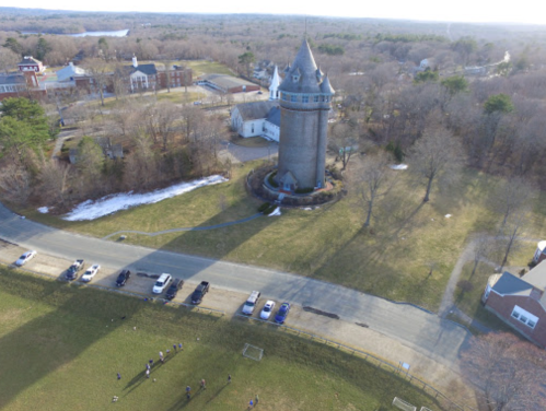 Aerial view of a stone tower surrounded by trees and a grassy area, with cars parked nearby and a few people in the distance.