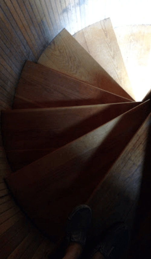 A spiral wooden staircase viewed from above, with a person's feet visible on the steps.