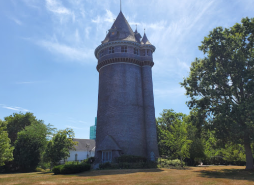 A tall, stone tower with a conical roof surrounded by greenery and a clear blue sky.