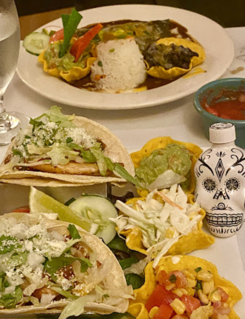 A colorful plate of tacos with toppings, guacamole, and a decorative skull bottle, alongside a rice and enchilada dish.