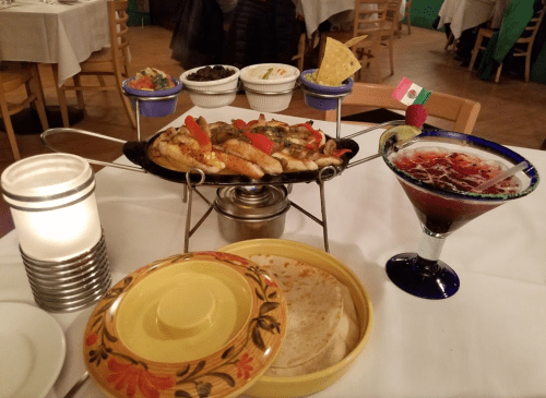 A vibrant Mexican meal featuring sizzling fajitas, tortillas, sides, and a colorful cocktail on a table.