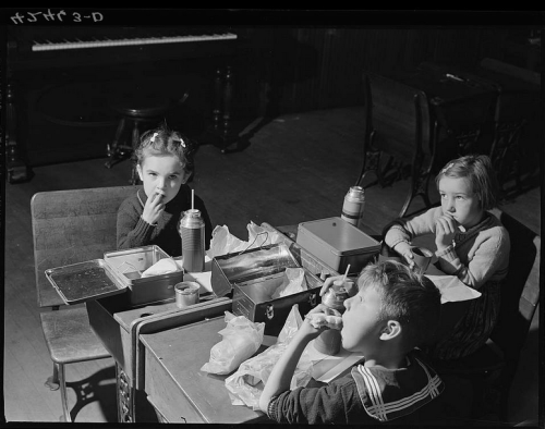 Three children sitting at a table, eating lunch from metal lunchboxes in a dimly lit room.