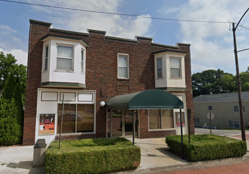 A brick building with two bay windows and a green awning, surrounded by low hedges and a sidewalk.