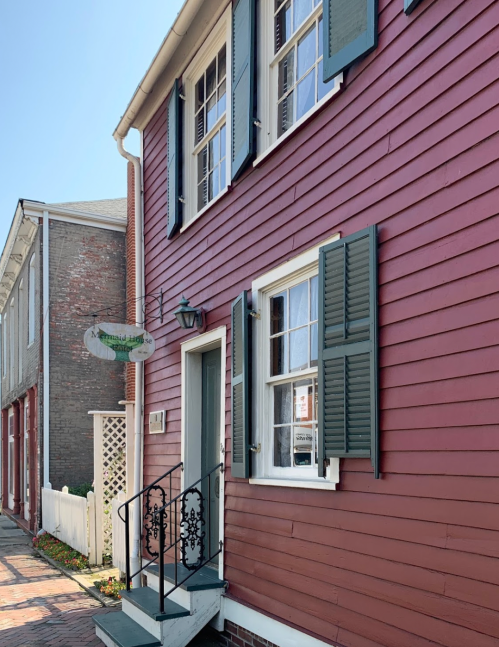 A charming red wooden house with green shutters and a decorative staircase, featuring a sign hanging nearby.
