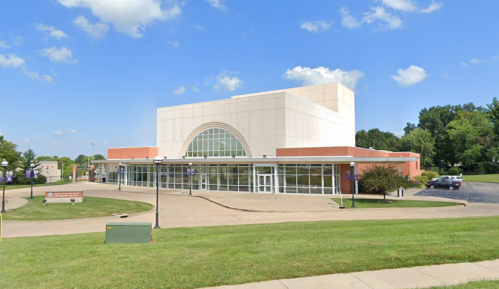 Modern building with large glass entrance, surrounded by green grass and trees under a blue sky with clouds.