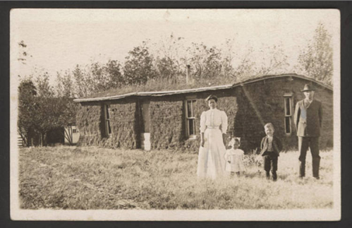 A vintage photo of a family standing outside a rustic house, with two children and adults in early 20th-century clothing.