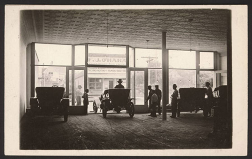 Historic interior of a car dealership with vintage cars and people, featuring large windows and a patterned ceiling.
