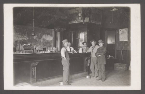 Black and white photo of four people at a bar, with a counter and vintage decor in a historic setting.
