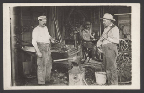 Three men in a workshop, one holding a tool, with an anvil and a wagon wheel in the background. Vintage black and white photo.