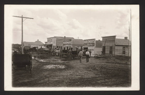 Historic black-and-white photo of a rural storefront with horse-drawn carriages and people in a dirt road setting.