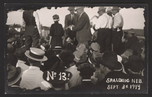 A historical black-and-white photo of a crowd in Spalding, Nebraska, on September 3, 1914, with men in hats and suits.