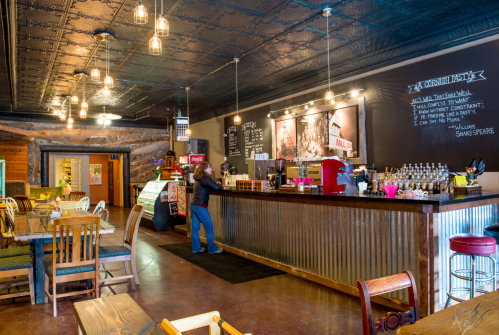A cozy café interior with a wooden bar, colorful decor, and a person standing at the counter. Warm lighting enhances the atmosphere.